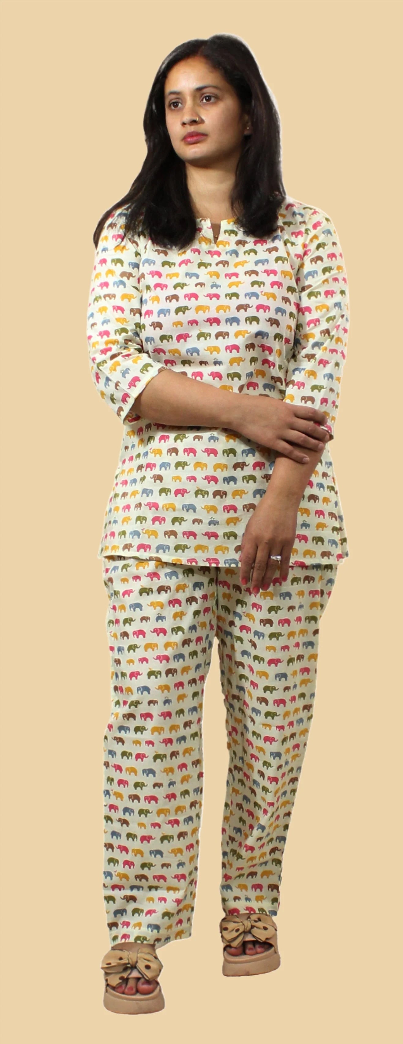 A woman wearing a Natural Fab night suit with a colorful elephant print. The night suit includes a long-sleeve top and matching pants. She is also wearing beige open-toe sandals with bows.